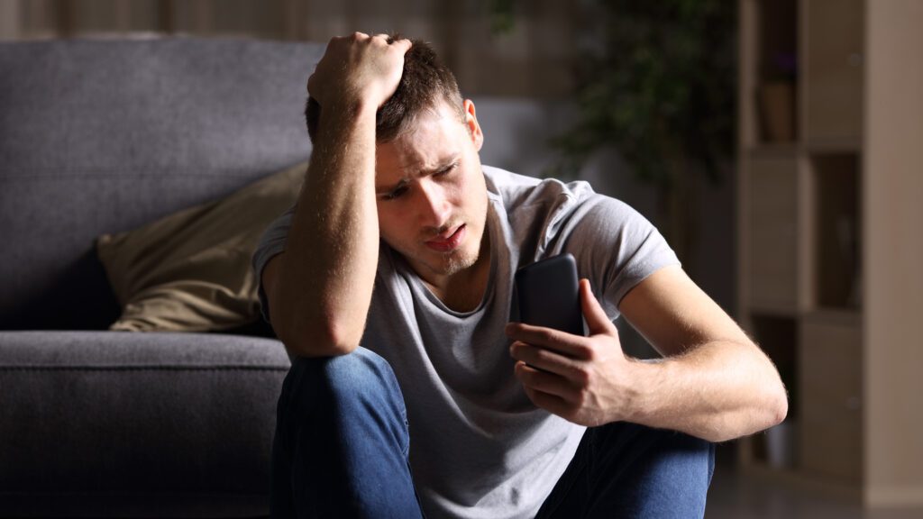 Single sad man checking mobile phone sitting on the floor in the living room at home.