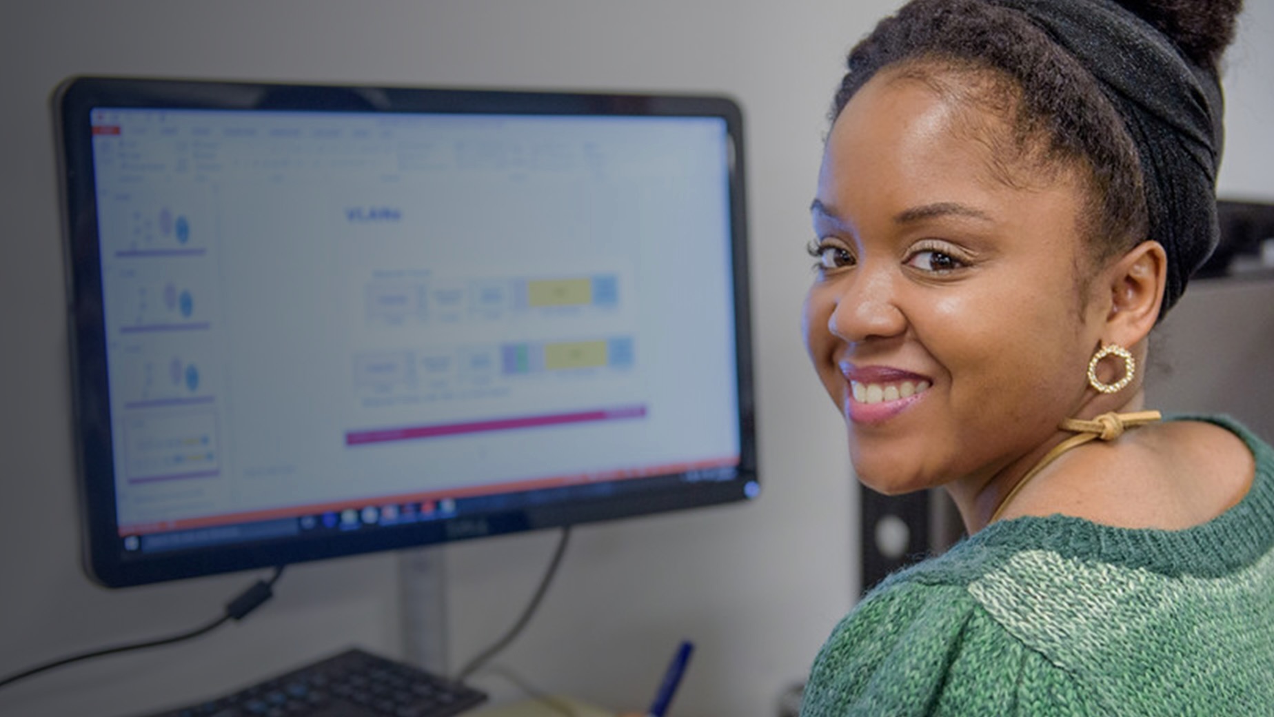 Smiling woman looking over her shoulder while sitting in front of desktop computer.