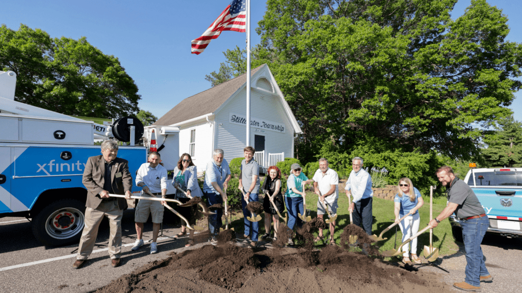 Members from Stillwater Township, Washington County, MInnesota Office of Broadband, and Comcast gathered in front of a pile of dirt with shovels to celebrate a groundbreaking ceremony. Every participant is holding a shovel and gently throwing dirt .