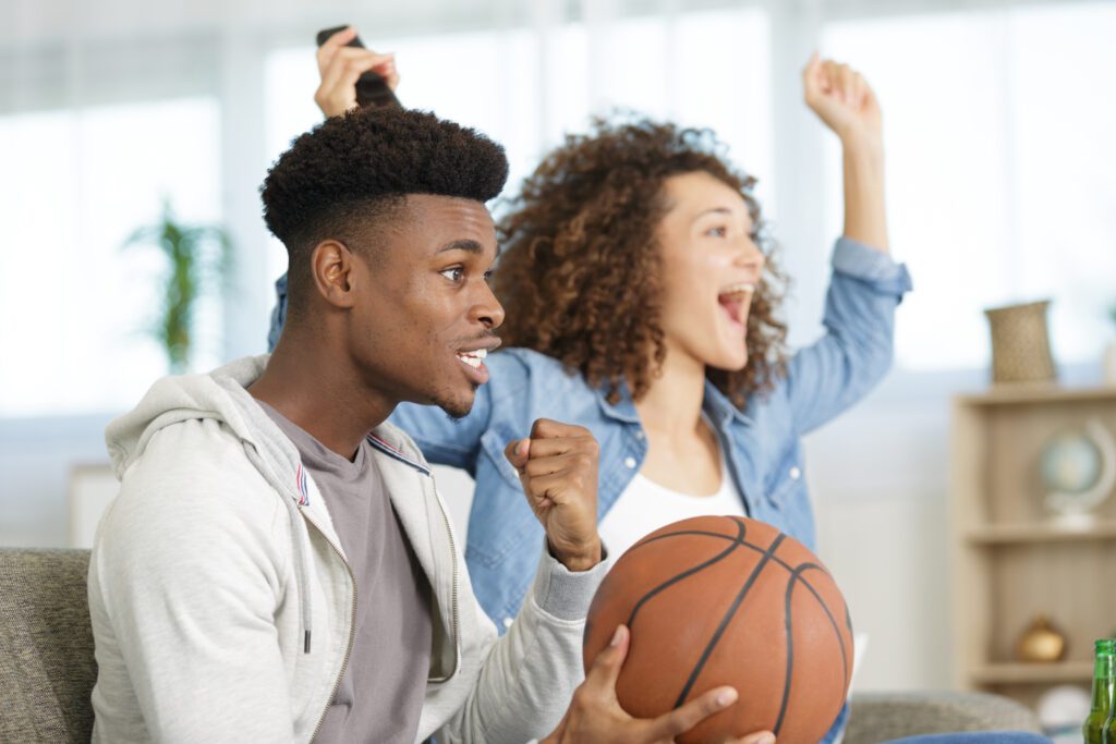 A male and female couple are watching basketball on a couch. March Madness fans are not only watching multiple games with Xfinity Multiview but keeping up with their March Madness brackets and scores only with Xfinity.