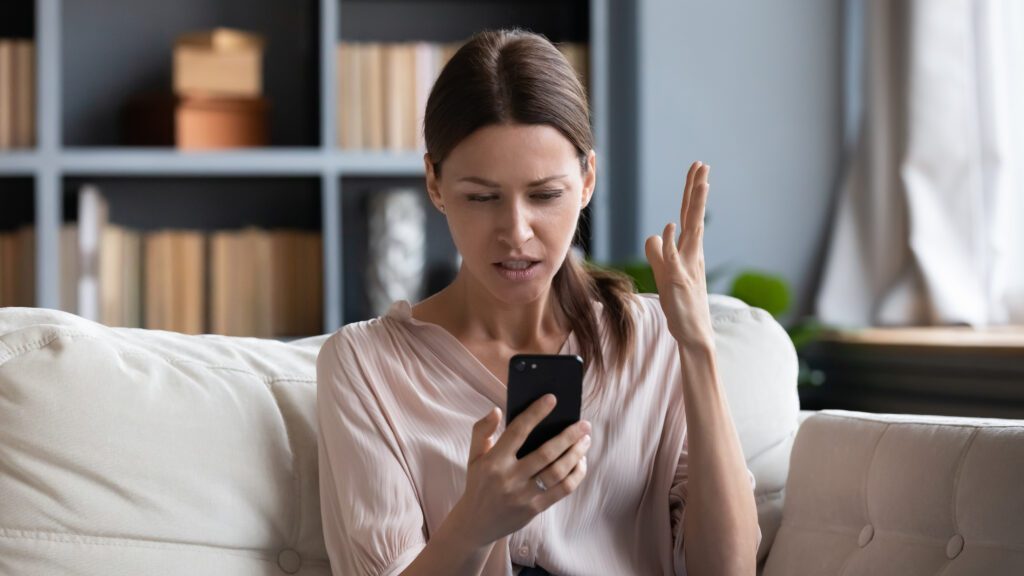 Confused angry woman having problem with phone, sitting on couch at home, unhappy young female looking at screen, dissatisfied by discharged or broken smartphone, reading bad news in message