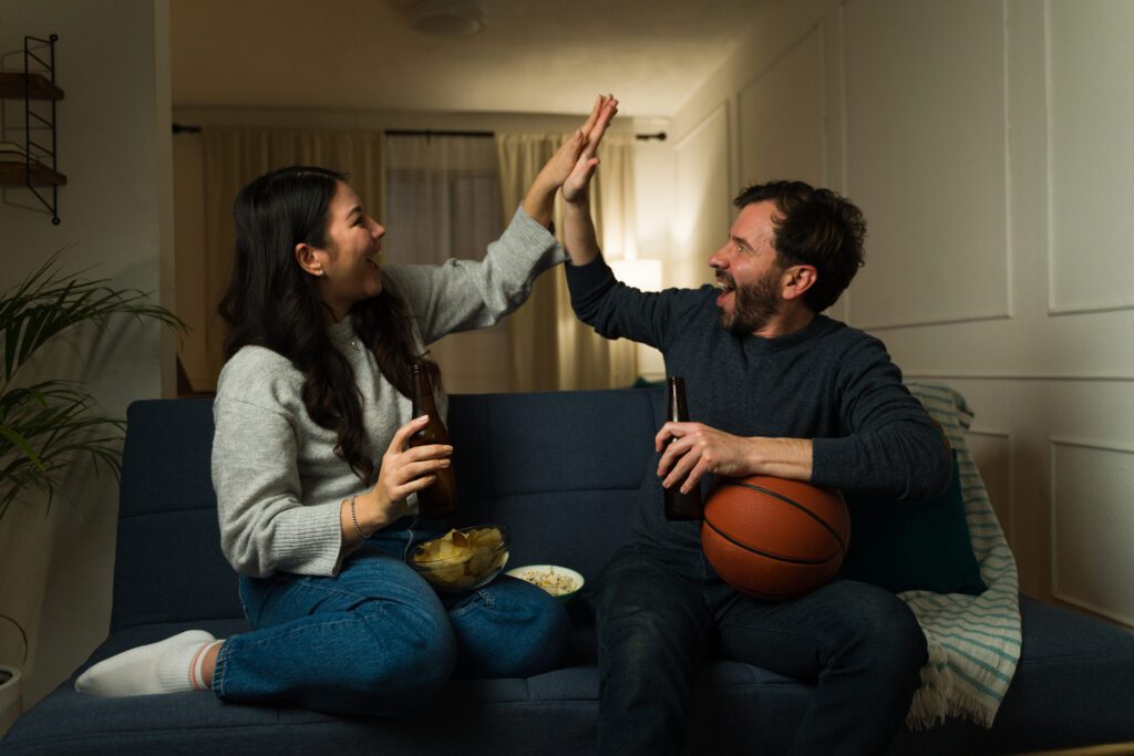 A male and female couple are watching basketball on a couch. and high fiving March Madness fans are not only watching multiple games with Xfinity Multiview but keeping up with their March Madness brackets and scores only with Xfinity.
