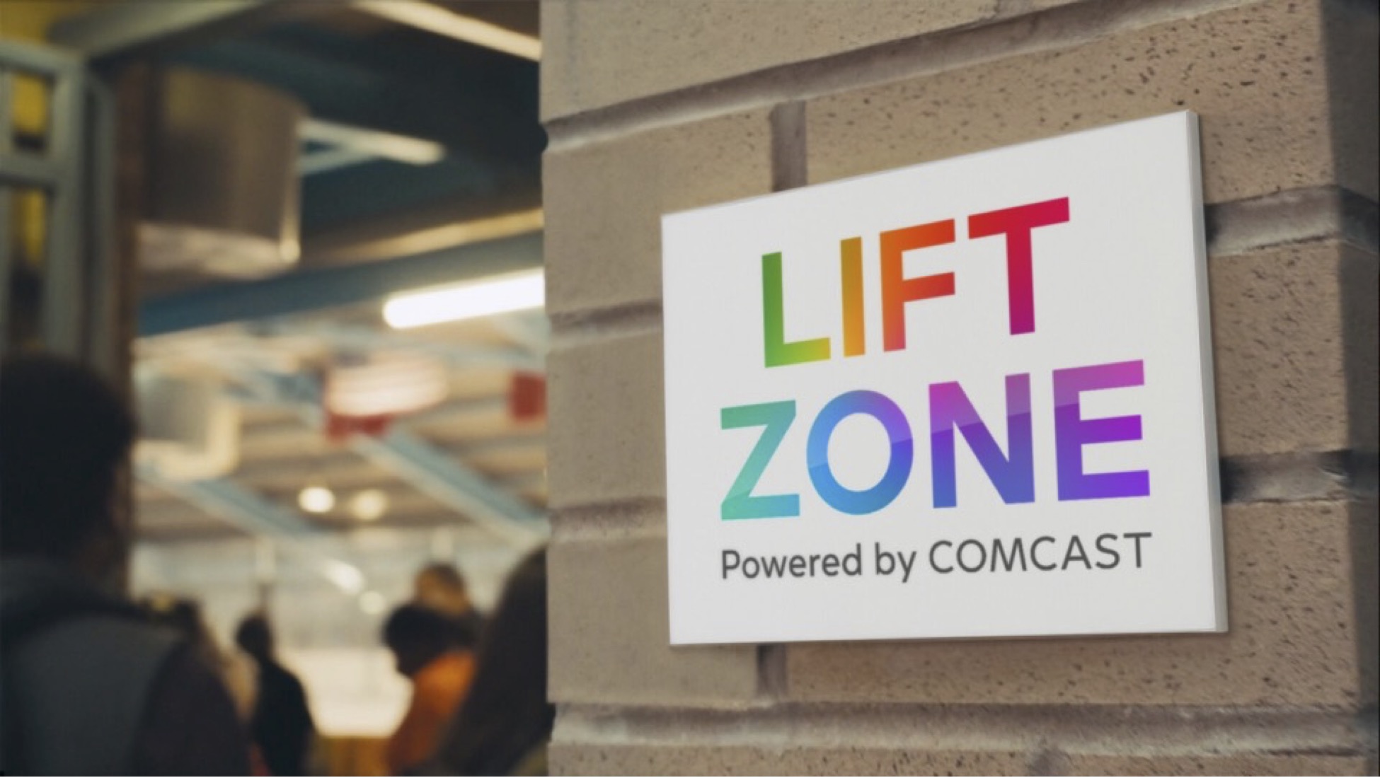 A white sign mounted on a beige brick wall reads 'LIFT ZONE' in colorful rainbow letters, with 'Powered by COMCAST' written below in gray. The sign is shown against a blurred background of what appears to be an indoor space with fluorescent lighting and people in the distance.